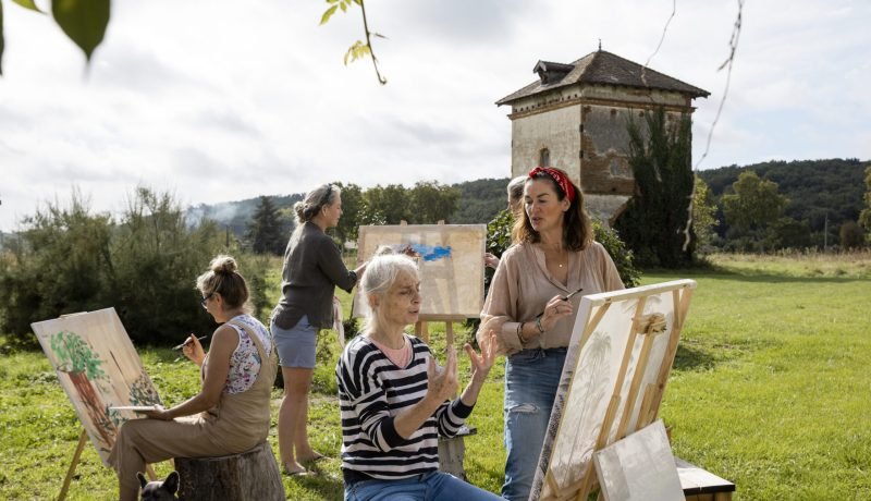 Mature women doing an art class in France while on a wellness retreat. They are painting on canvas together sitting outdoors with beautiful scenery around them with the help of art tutors.