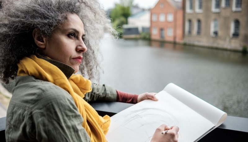 Mature woman artist making a drawing over a canal in London. She is wearing a military jacket and yellow scarf.